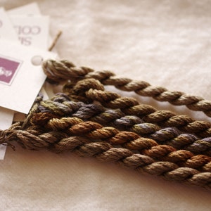 May include: Four skeins of embroidery floss in shades of brown, green, and purple. The skeins are labeled "100% Silk" and "Cobweb" with a small logo.