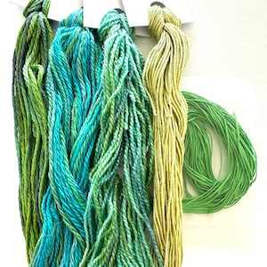 May include: Close-up of several skeins of colorful embroidery floss. The floss is in shades of green, teal, and yellow-green. The skeins are arranged vertically, with a single, tightly wound green skein at the bottom right. The floss appears soft and textured.