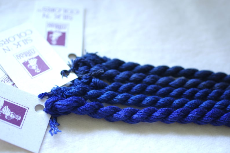 May include: A collection of six skeins of blue silk embroidery floss. The floss is arranged in a fan shape and is ready for use in a craft project.