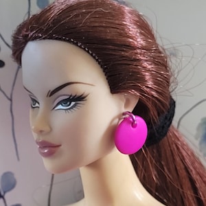 May include: A doll with long brown hair wearing a pink dress and bright pink round earrings.