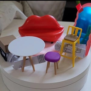 May include: A miniature furniture set featuring a bright red lip-shaped sofa, a white circular table with three light wood legs, a yellow chair with a tan seat, a small purple stool, a tan cabinet, and a tall blue mirror with a pink wavy frame.