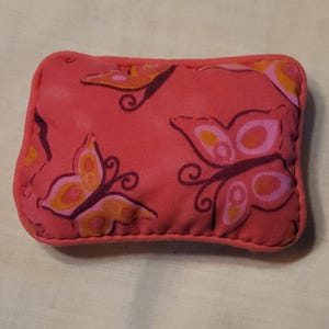 May include: A rectangular fabric pin cushion with rounded corners, featuring a coral pink background printed with stylized butterflies in shades of orange, dark purple, and light pink. The edges are finished with a matching coral pink fabric border.