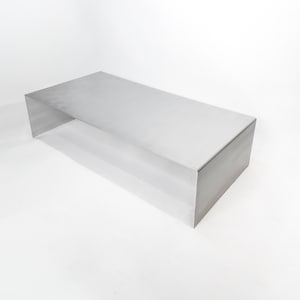 Könnte beinhalten: Ein rechteckiger Couchtisch aus gebürstetem Metall. Der Tisch ist eine solide, kastenartige Struktur mit flacher Oberseite und Seiten. Das Metall hat eine matte Oberfläche, die das Licht reflektiert. Der Tisch steht vor einem weißen Hintergrund.