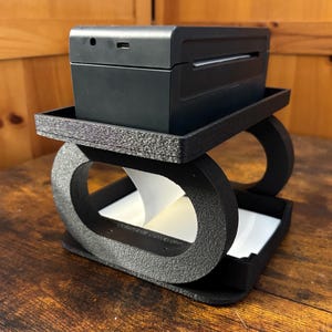 May include: A black thermal printer sits atop a textured black stand with an oval design. The stand has a tray for paper and a lower tray. The printer is rectangular with a paper output slot.