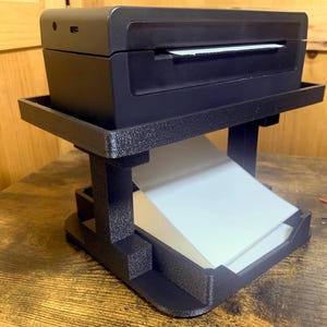 May include: A black printer with a white paper tray sits on a black 3D printed stand. The stand has a small shelf for storing extra paper.