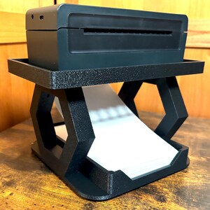 May include: A black 3D printed stand with a hexagonal design holds a black photo printer. The stand has a built-in tray for holding photos.