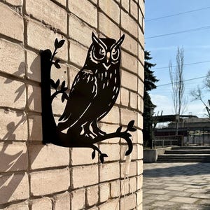 May include: A black metal owl silhouette decoratively mounted on a brick wall. The owl is perched on a branch. A white hanging planter with red flowers hangs below. The scene is outdoors on a sunny day.
