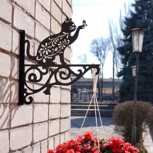 May include: Black metal cat-shaped plant hanger mounted on a brick wall. The cat is reaching for a butterfly. A white pot filled with red flowers hangs below. The brick wall is light-colored.
