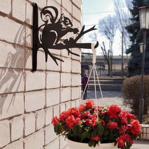 Könnte beinhalten: Schwarzer Metallhalter in Eichhörnchenform an einer Ziegelmauer, der einen weißen Hängepflanzentopf mit roten Blumen hält. Die Halterung zeigt ein Eichhörnchen auf einem Ast.
