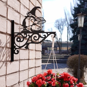 May include: A black metal hanging bracket with a gnome design, mounted on a brick wall. A white hanging planter filled with vibrant red flowers is suspended from the bracket. The scene is outdoors, with a sunny day and a paved walkway.