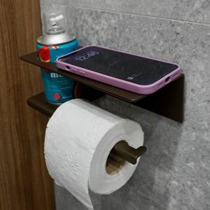May include: A modern bathroom shelf with a roll of toilet paper, a blue and silver spray can, and a smartphone with a pink case. The shelf is mounted on a gray tiled wall, with a wooden door in the background.