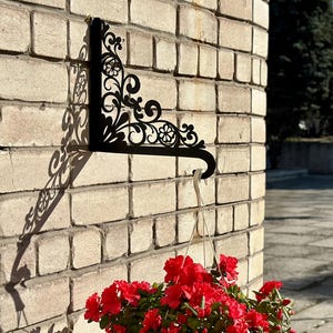 May include: A black, ornate metal bracket is mounted on a brick wall, casting a decorative shadow. A white hanging planter filled with vibrant red flowers is suspended from the bracket, creating a striking contrast against the brick.