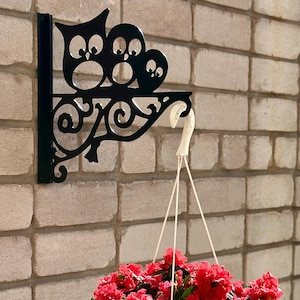May include: Black metal owl-themed hanging bracket mounted on a brick wall. The bracket supports a white hanging planter filled with vibrant red flowers. The bracket features three owl silhouettes and decorative scrollwork.