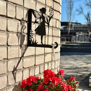 Könnte beinhalten: Schwarze Metall-Wandkunst mit einer Feen-Silhouette, die Blumen hält, an einer Backsteinmauer befestigt. Ein weißer Blumentopf mit leuchtend roten Blumen hängt darunter und schafft eine dekorative Gartenszene.