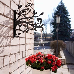 Puede incluir: Soporte de pared de metal negro con diseño de pájaro y rama. Una maceta colgante blanca llena de flores rojas está suspendida del soporte. El soporte está montado en una pared de ladrillos blancos.