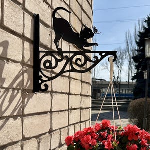 May include: Black metal cat-shaped plant hanger with a butterfly detail, mounted on a brick wall. The cat is reaching for the butterfly. A white hanging planter with red flowers is suspended below the bracket.