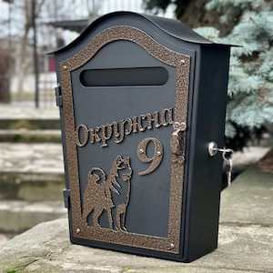 Puede incluir: Buzón de metal negro con un marco y detalles decorativos de color bronce. El buzón tiene una ranura para el correo, una cerradura con llaves y un diseño decorativo de dos perros y el número 9. La palabra "Окружна" está escrita.