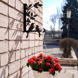 Puede incluir: Arte de pared de metal negro con dos pájaros posados en una rama con hojas. Una maceta colgante blanca, llena de flores rojas, cuelga debajo, contra una pared de ladrillos.