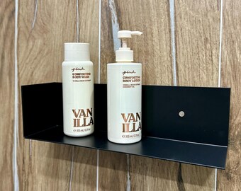 Puede incluir: Dos botellas blancas de gel de ducha y loción corporal con el texto "VANILLA" en color cobre, sobre una estantería de metal negro. La estantería está montada en una pared de azulejos de madera. Las botellas están etiquetadas como "Comforting Body Wash" y "Comforting Body Lotion".