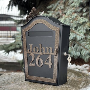 Puede incluir: Un buzón de metal negro con una mariposa decorativa en la parte superior. El buzón tiene un borde de color bronce y el nombre "John" y el número "264" se muestran en la parte delantera. Una llave está en la cerradura.