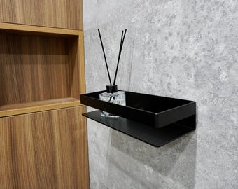 Puede incluir: Estante de pared de metal negro con una bandeja rectangular y un estante inferior. Un frasco difusor de vidrio transparente con cañas negras se encuentra en la bandeja. El estante está montado en una pared de azulejos grises.