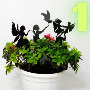 Könnte beinhalten: Schwarze Metall-Feengartenstecker in einem weißen Topf mit grünem Laub und rosa Blüten. Die Stecker zeigen Feen und einen Vogel. Die Zahl 1 befindet sich im Hintergrund.