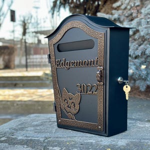 May include: A black mailbox with a decorative bronze-colored frame and accents. The mailbox features the name "Edgemont" and the number "3022". A key hangs in the lock, and a cat silhouette is visible on the front.