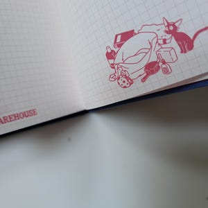 May include: Open notebook with a grid pattern on the pages. A red illustration of a cat and a pile of items, including a bag, bottle, and brush, is in the corner. The word "WAREHOUSE" is printed in red along the edge.
