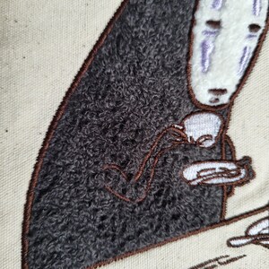 May include: Embroidered image of a character from Spirited Away, holding a teacup and saucer. The character has a white face with purple markings and a dark gray, textured body. The embroidery is on a cream-colored fabric, with brown outlining.