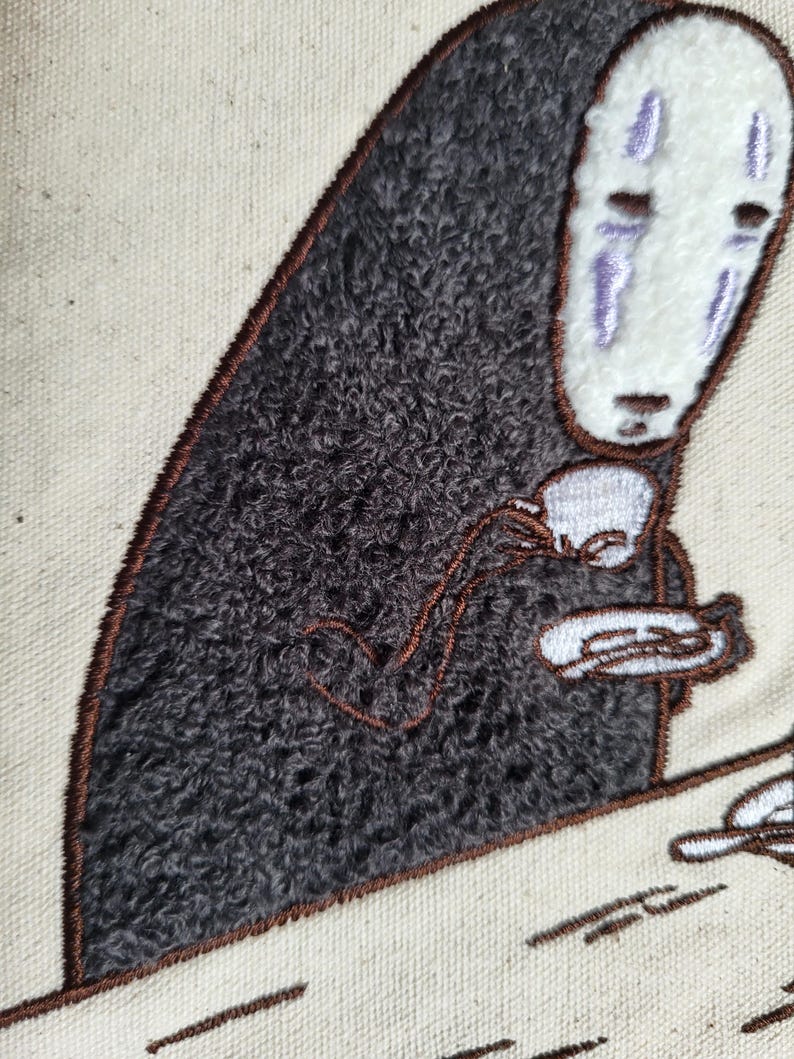 May include: Embroidered image of a character from Spirited Away, holding a teacup and saucer. The character has a white face with purple markings and a dark gray, textured body. The embroidery is on a cream-colored fabric, with brown outlining.