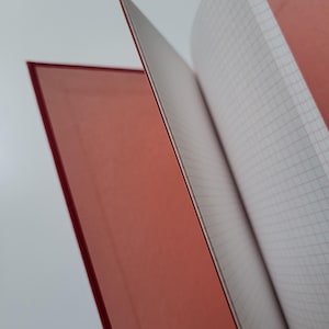 May include: A red notebook with a grid-lined interior. The pages are open, revealing the white paper with a red grid. The cover is a solid, matte red. The notebook is ideal for note-taking or journaling.