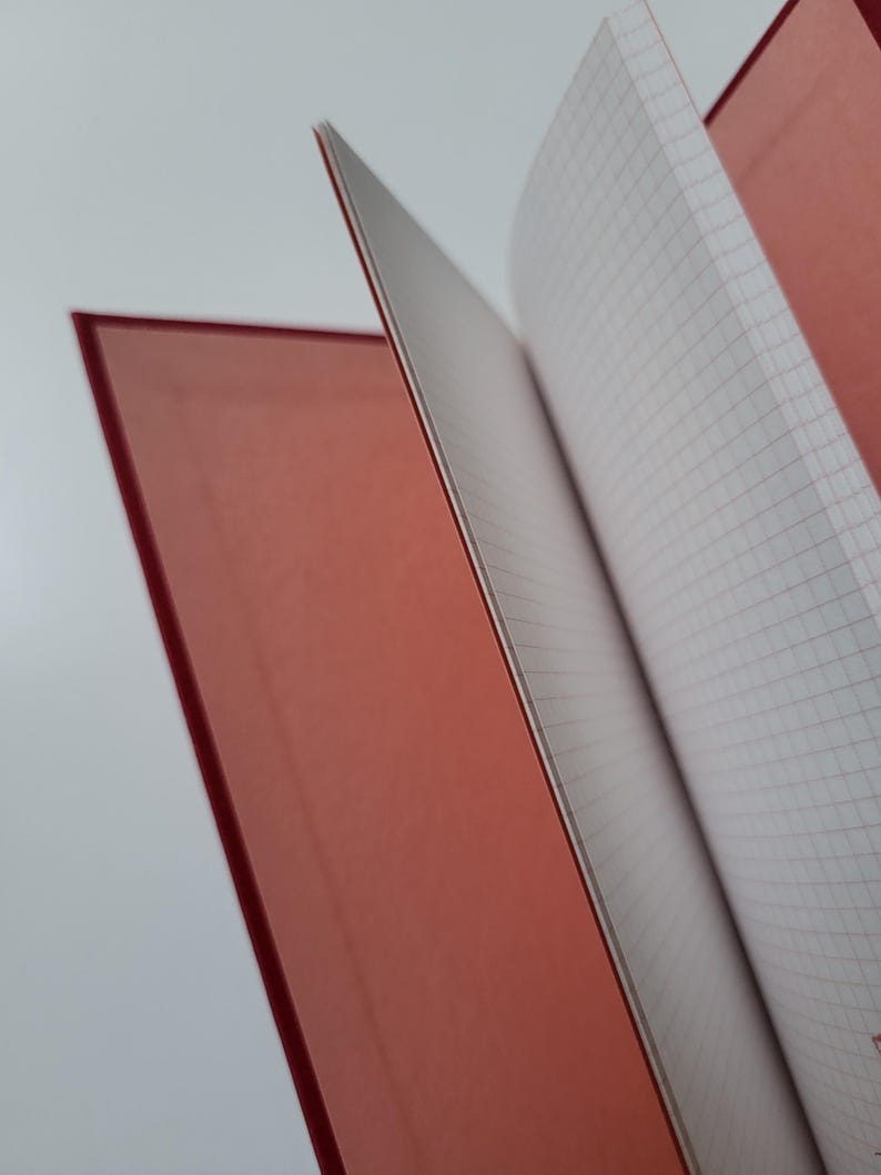 May include: A red notebook with a grid-lined interior. The pages are open, revealing the white paper with a red grid. The cover is a solid, matte red. The notebook is ideal for note-taking or journaling.