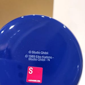 May include: A close-up of a glossy, cobalt blue object with white specks. The object has text that reads "Studio Ghibli" and "1989 Eiko Kadono - Studio Ghibli - N". A red square with a white "S" is also visible.