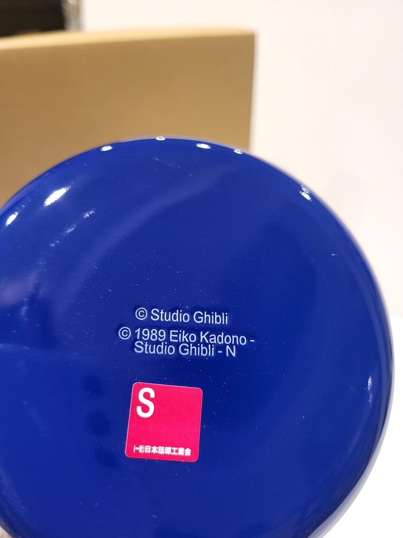 May include: A close-up of a glossy, cobalt blue object with white specks. The object has text that reads "Studio Ghibli" and "1989 Eiko Kadono - Studio Ghibli - N". A red square with a white "S" is also visible.