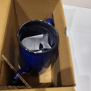 May include: A blue enamel mug with a handle, inside a cardboard box. The mug has a dark blue exterior and a black rim. The handle is also blue. The mug appears to be new, with a piece of paper inside.
