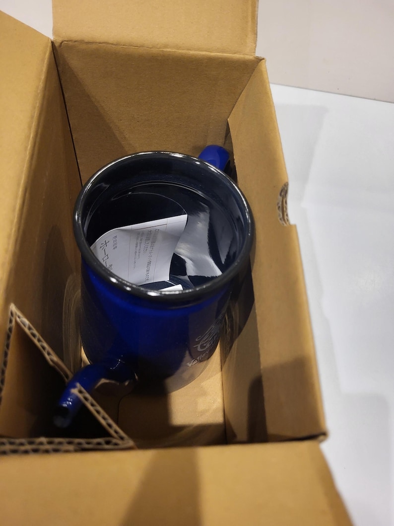 May include: A blue enamel mug with a handle, inside a cardboard box. The mug has a dark blue exterior and a black rim. The handle is also blue. The mug appears to be new, with a piece of paper inside.