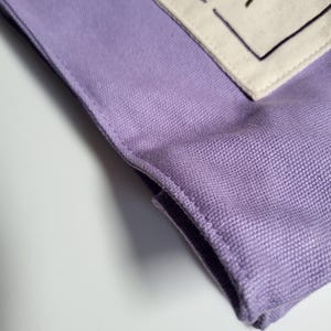 May include: Close-up of a lavender fabric item with a textured weave. A section of off-white fabric with a dark gray shape and black stitching is visible. The item has a folded edge and is set against a white background.