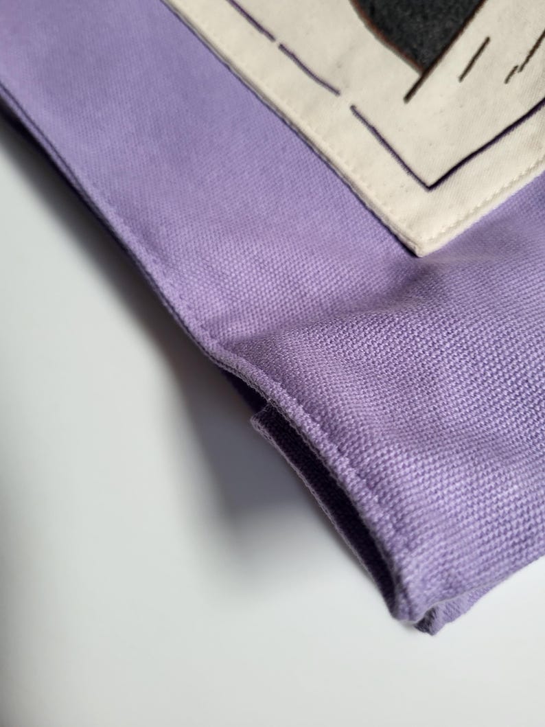 May include: Close-up of a lavender fabric item with a textured weave. A section of off-white fabric with a dark gray shape and black stitching is visible. The item has a folded edge and is set against a white background.