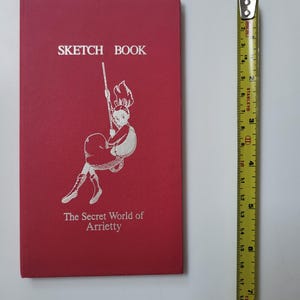 May include: A red sketchbook with the text "SKETCH BOOK" at the top and "The Secret World of Arrietty" at the bottom. The cover features a white illustration of a girl swinging. A yellow tape measure is next to the book.