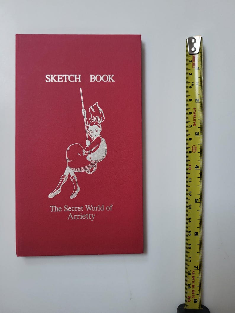 May include: A red sketchbook with the text "SKETCH BOOK" at the top and "The Secret World of Arrietty" at the bottom. The cover features a white illustration of a girl swinging. A yellow tape measure is next to the book.