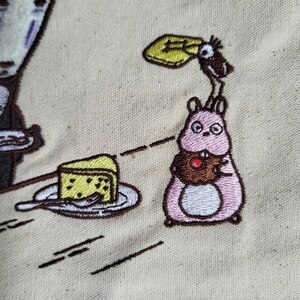May include: Embroidered design featuring characters from the animated film Spirited Away. A pink creature holds a cupcake, a slice of cake sits on a plate, and a faceless spirit is also present. The design is on a beige fabric.