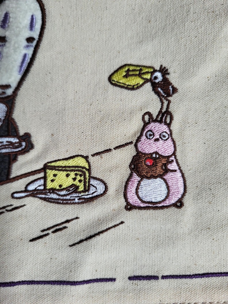 May include: Embroidered design featuring characters from the animated film Spirited Away. A pink creature holds a cupcake, a slice of cake sits on a plate, and a faceless spirit is also present. The design is on a beige fabric.