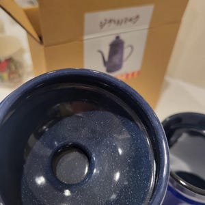 May include: Close-up of a dark blue ceramic coffee pot with a speckled finish. The pot has a wide opening and a small hole in the center. A cardboard box with Japanese text and a drawing of a coffee pot is in the background.