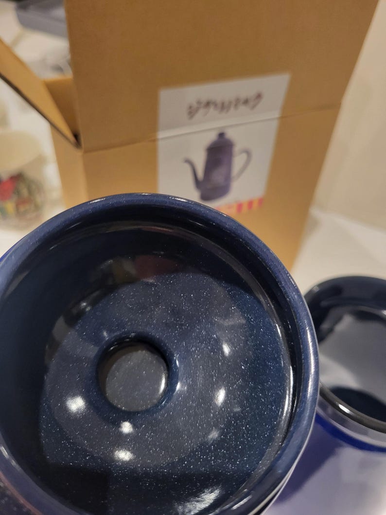 May include: Close-up of a dark blue ceramic coffee pot with a speckled finish. The pot has a wide opening and a small hole in the center. A cardboard box with Japanese text and a drawing of a coffee pot is in the background.