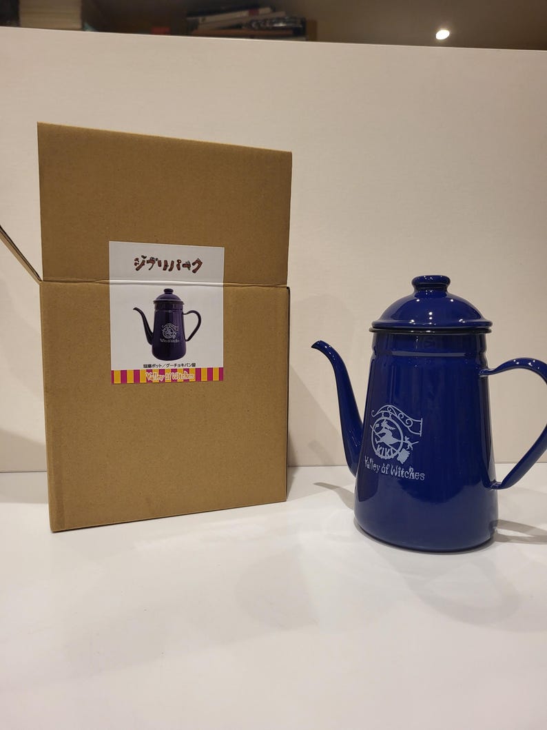 May include: A cobalt blue enamel kettle with a spout and handle, next to a cardboard box. The kettle has a logo that says "Valley of Witches". The box has Japanese text and a picture of the kettle.
