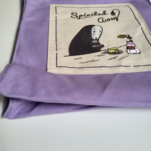 May include: A lavender tote bag featuring an embroidered design from the movie "Spirited Away." The design includes the text "Spirited Away" and a character with a white mask, a piece of cake, and a small creature. The bag has two handles.