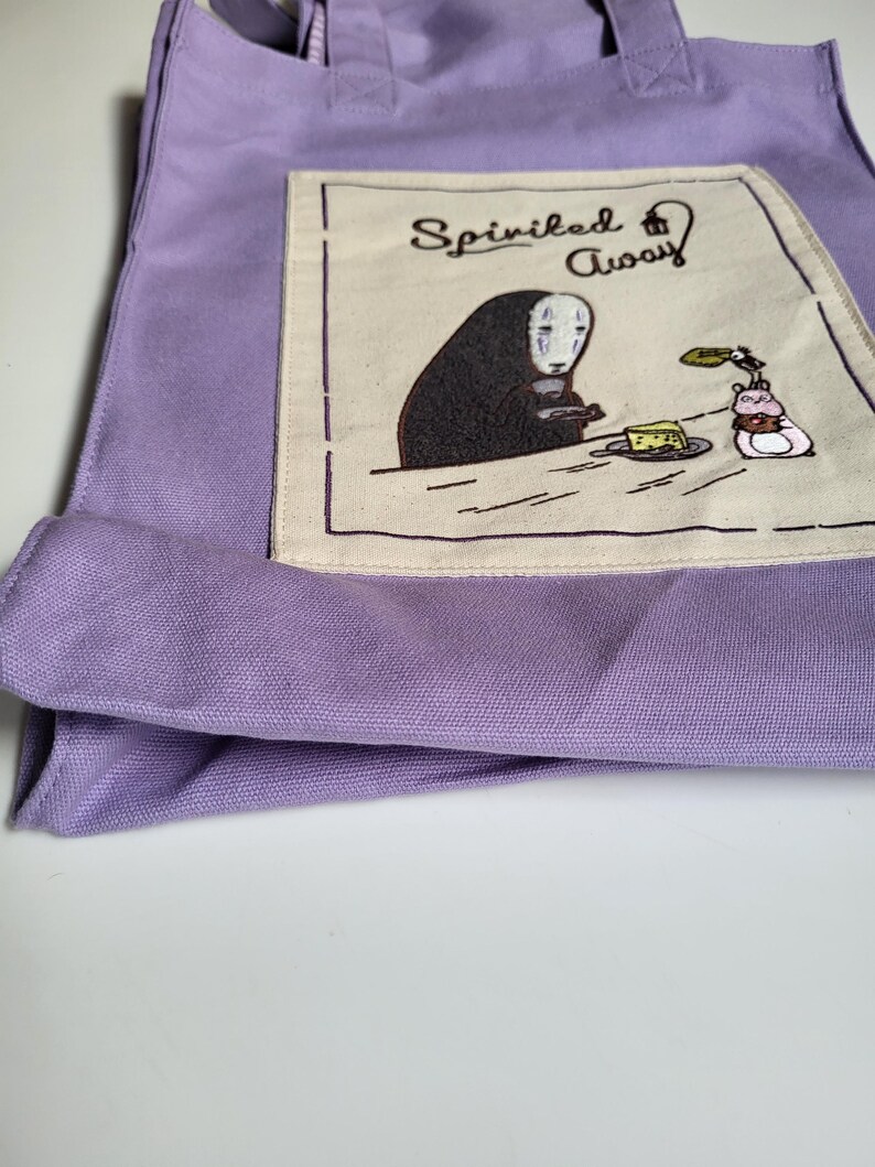 May include: A lavender tote bag featuring an embroidered design from the movie "Spirited Away." The design includes the text "Spirited Away" and a character with a white mask, a piece of cake, and a small creature. The bag has two handles.