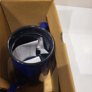 May include: A cobalt blue enamel mug with a black rim and a matching blue handle. The mug is inside a brown cardboard box, suggesting it's new and packaged for shipping. The mug appears to be empty, with a white paper inside.