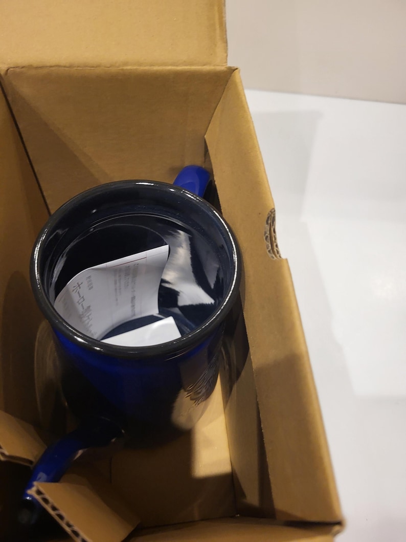 May include: A cobalt blue enamel mug with a black rim and a matching blue handle. The mug is inside a brown cardboard box, suggesting it's new and packaged for shipping. The mug appears to be empty, with a white paper inside.