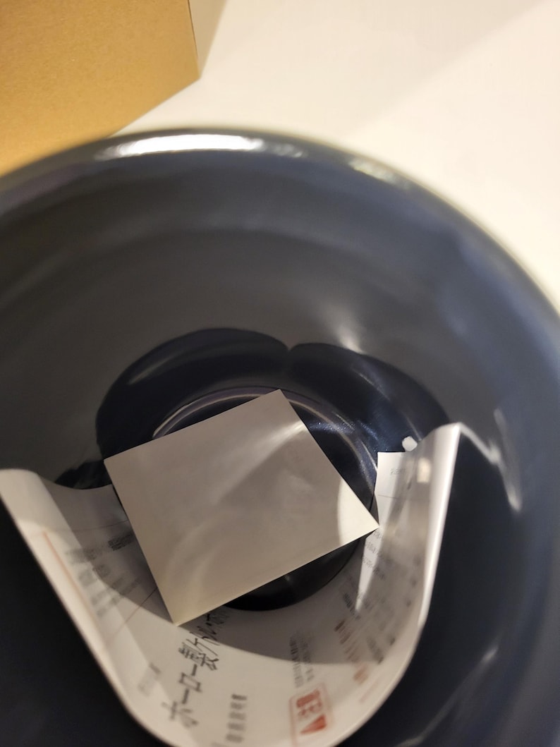 May include: A dark blue, glossy ceramic bowl with a white square card and a partially visible paper with Japanese text inside. The bowl's interior reflects light, highlighting its smooth surface.